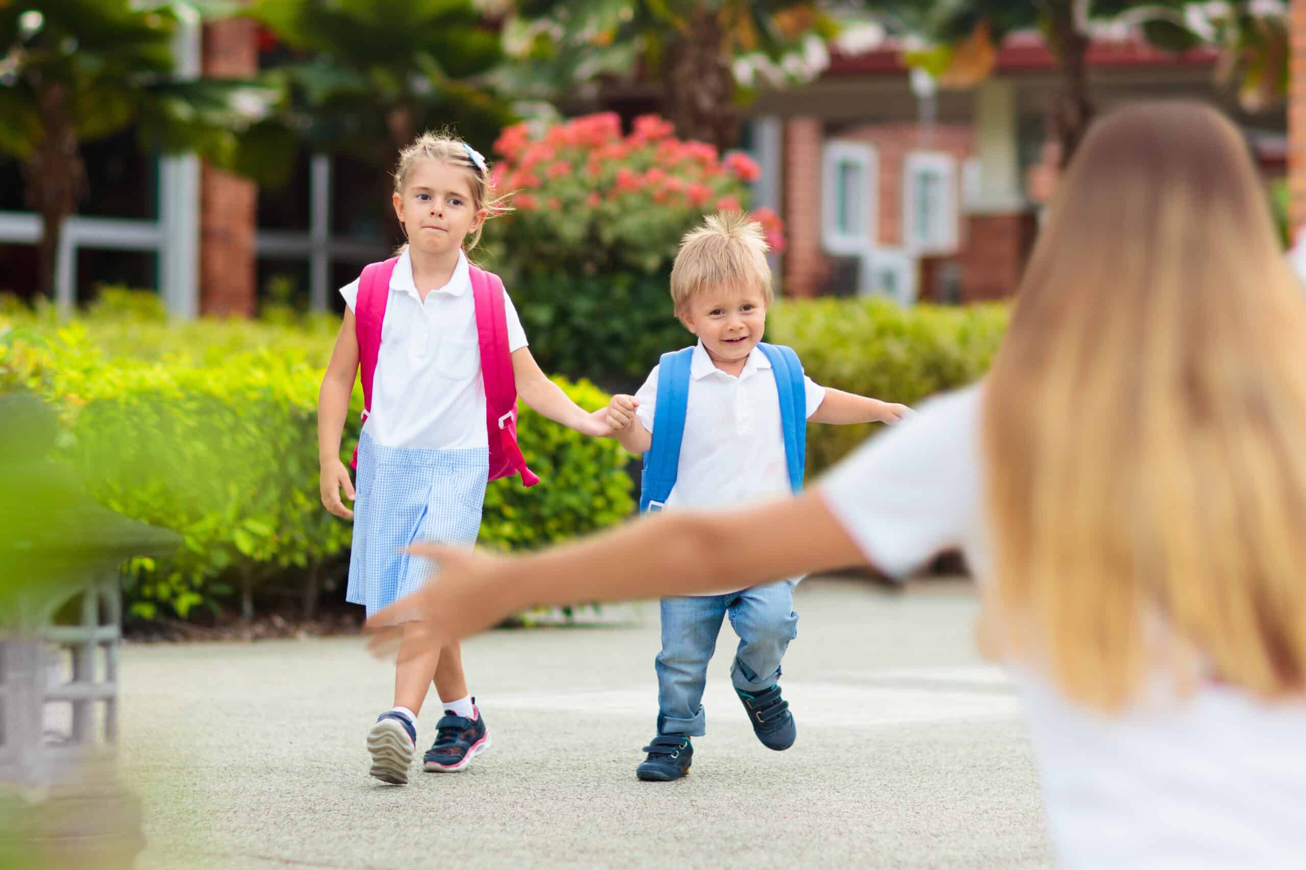 Young children wearing backpacks leaving school and walking towards a woman with outstretched arms.